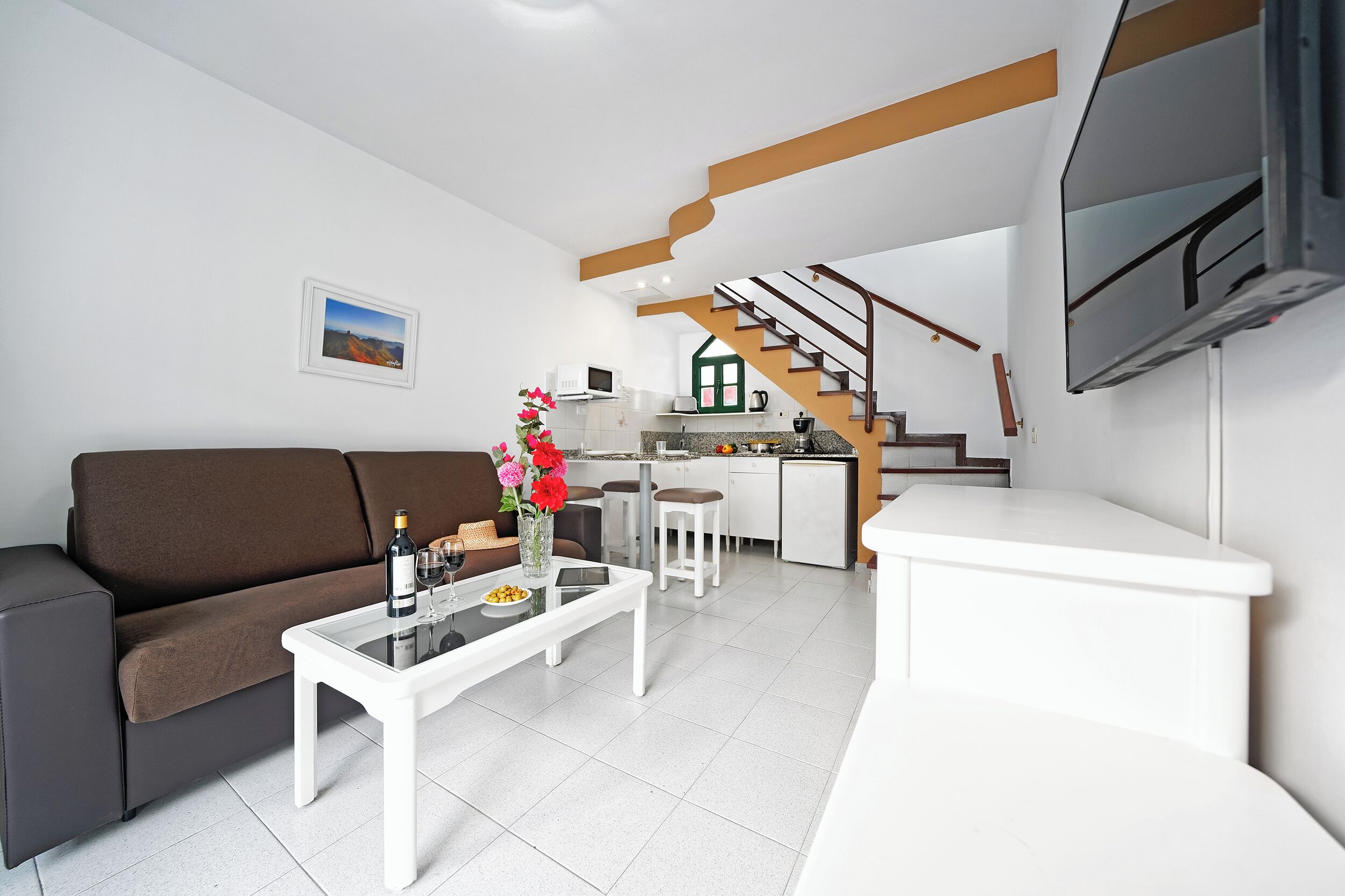 A modern living room with a sofa, coffee table, and a TV. A kitchen with a staircase is visible in the background.