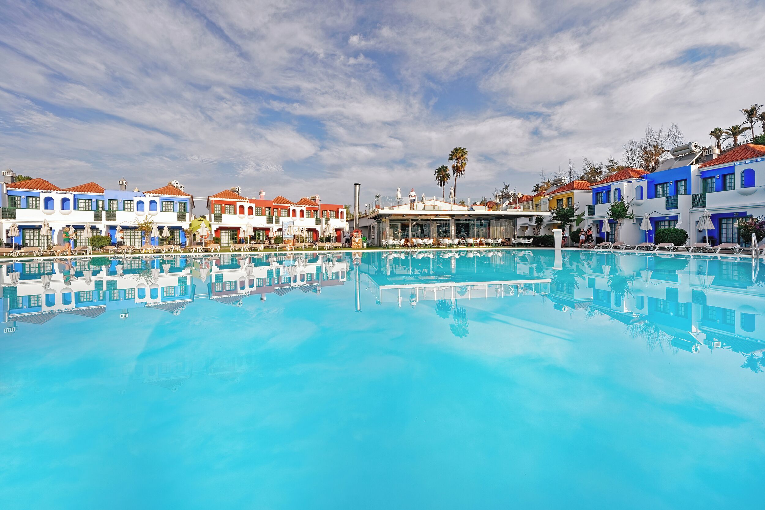 Una piscina que refleja edificios coloridos, un cielo despejado y palmeras.