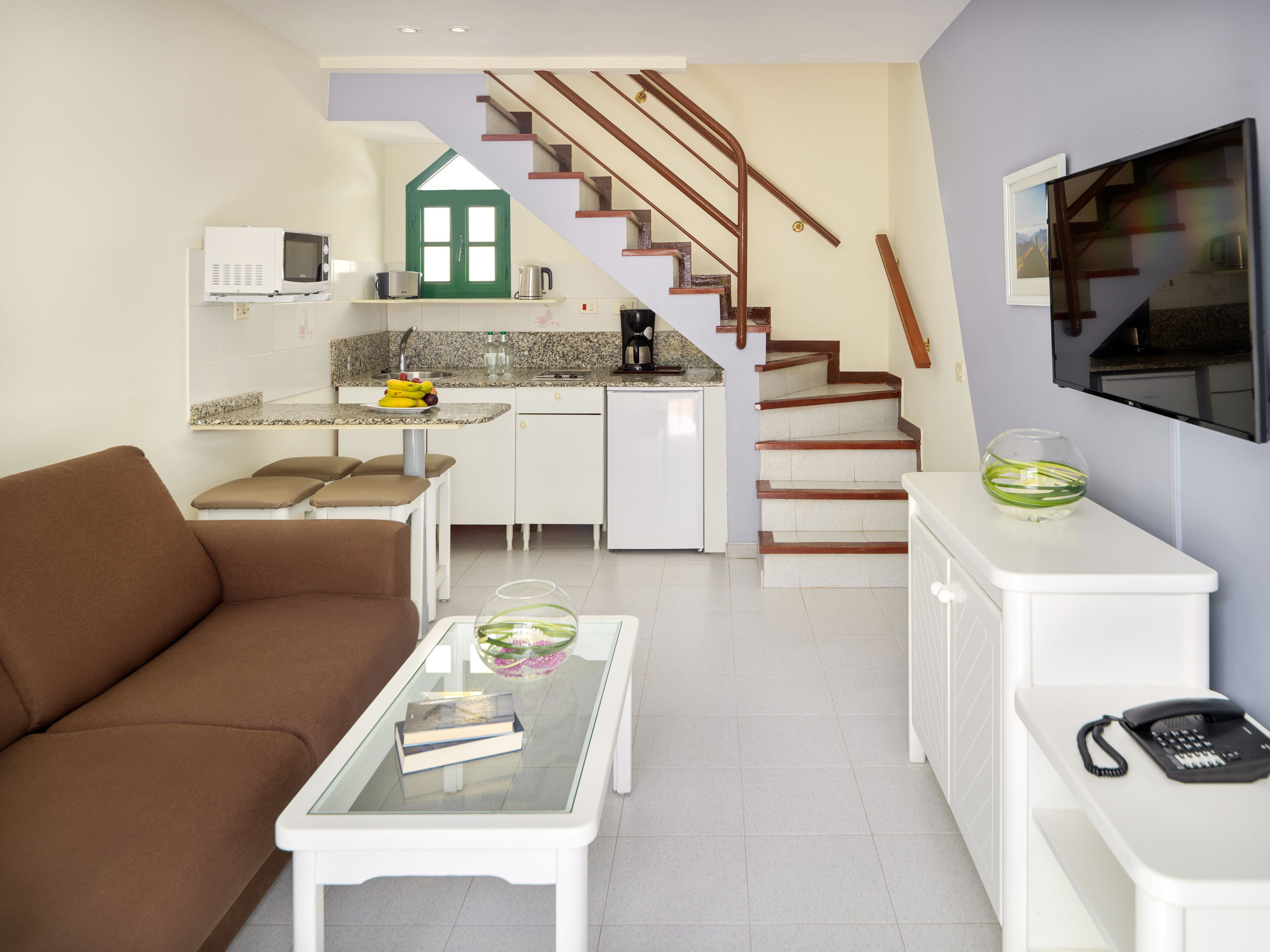 Ein kompakter Wohnraum mit Küchenzeile, einem braunen Sofa, einem Glastisch und einer Treppe, die in ein Obergeschoss führt.
