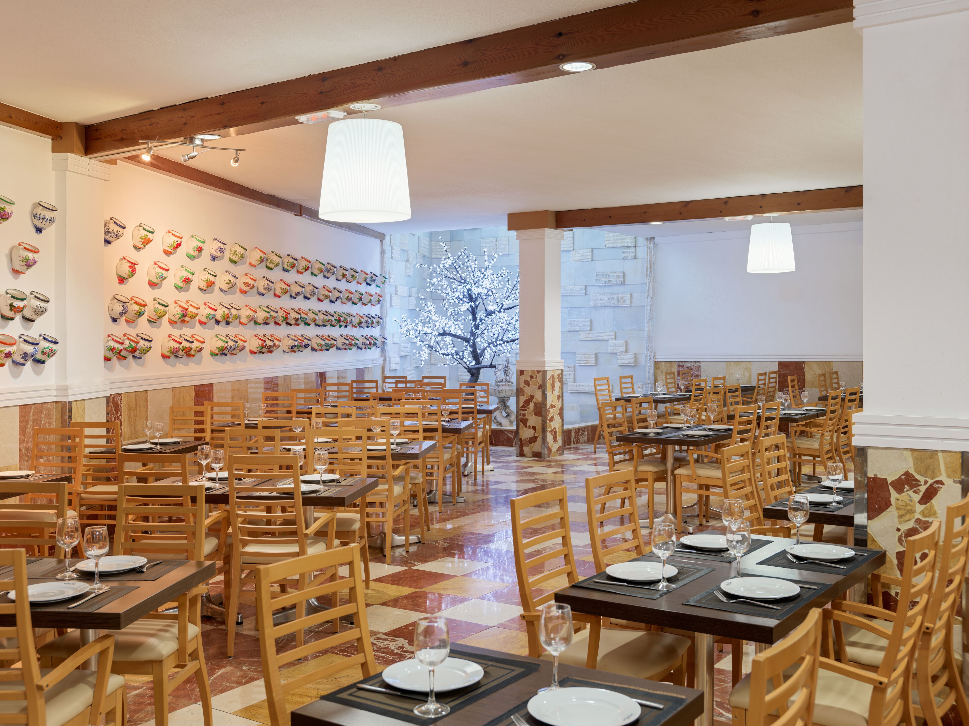 Un comedor con varias mesas y sillas, una pared decorada con macetas de cerámica y un gran árbol con flores blancas.