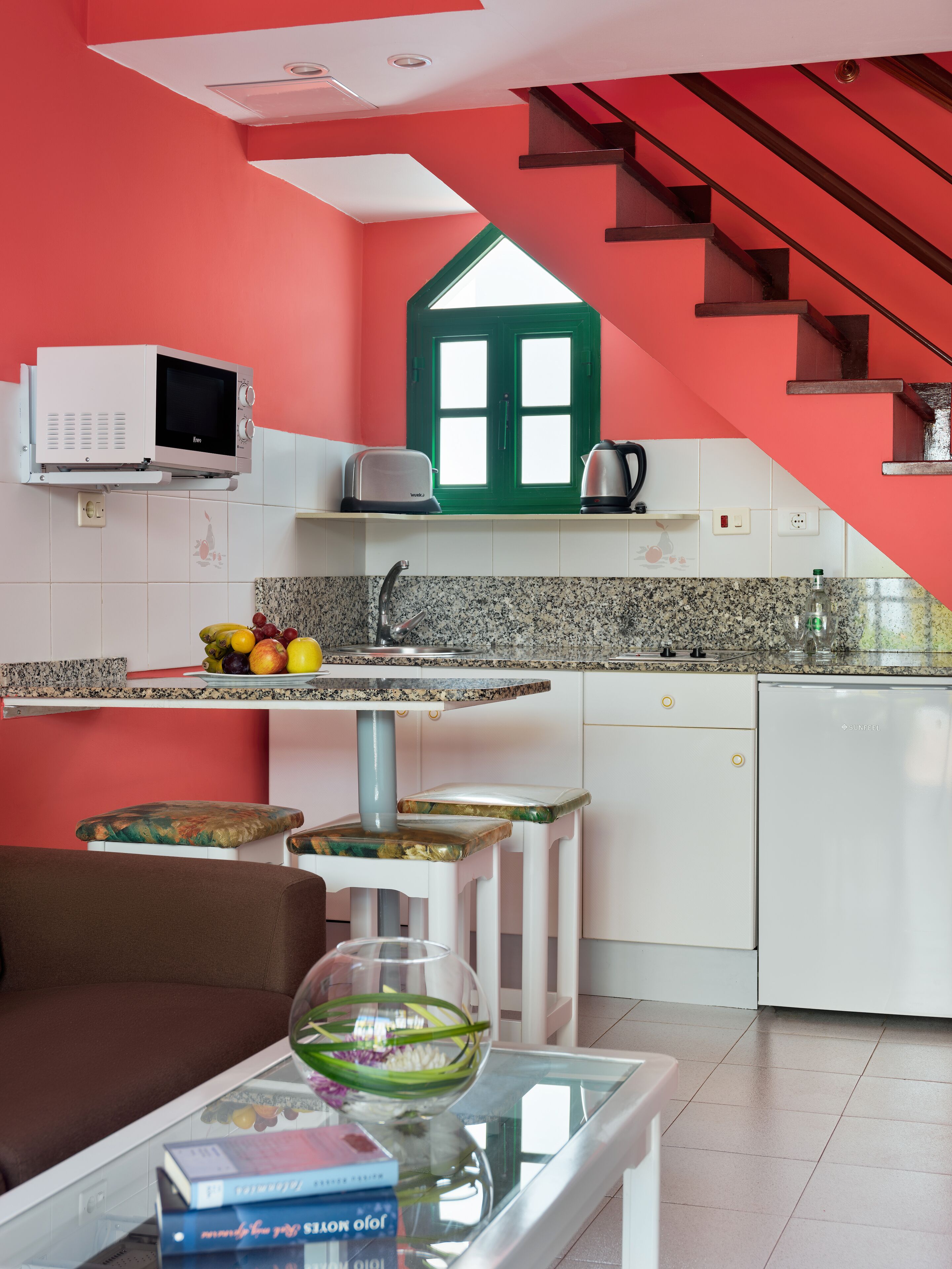 Eine moderne Küche mit Mikrowelle, Toaster und Wasserkocher auf der Arbeitsfläche, ein Essbereich mit einem Glastisch und Büchern sowie eine Treppe mit einer grünen Tür.