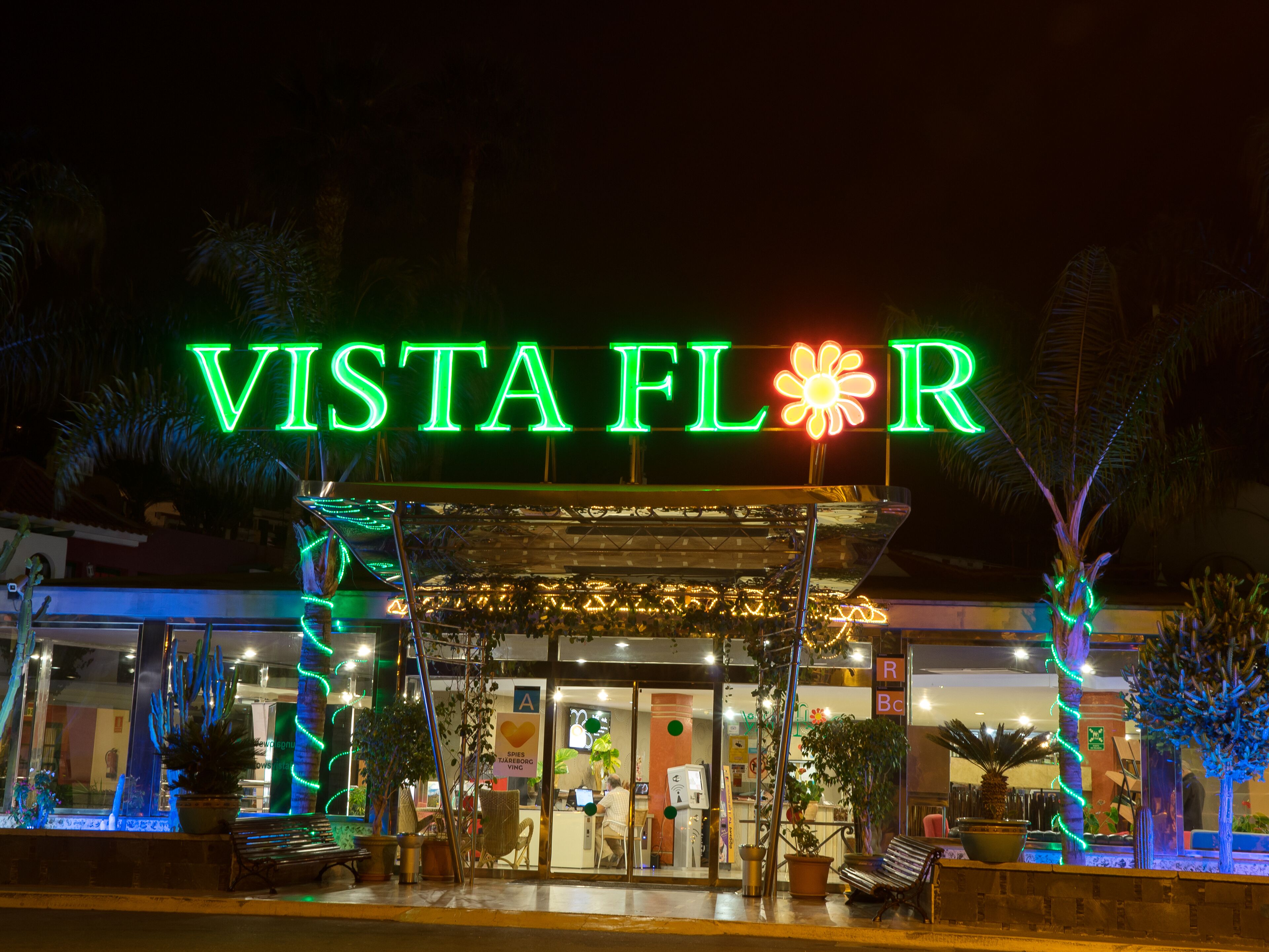 A hotel entrance with a neon sign reading "VISTA FLOR" and a covered outdoor seating area with potted plants.