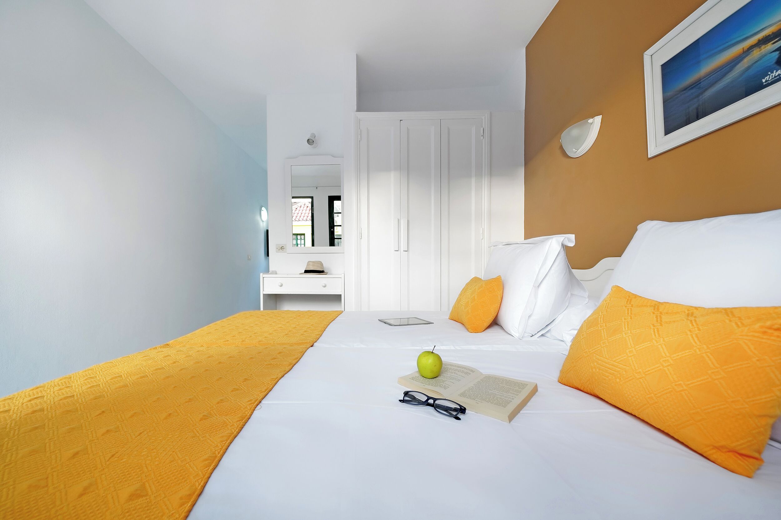 Una habitación de hotel con cama, mesita de noche, espejo, puerta y un cuadro en la pared.