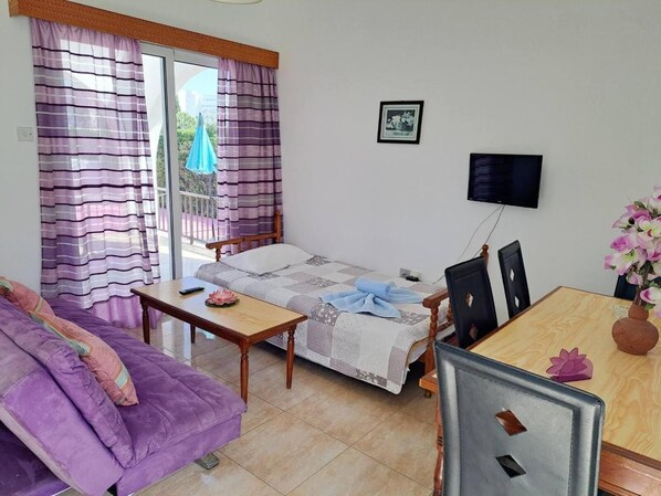 A small, tidy room with a bed, a sofa, a dining table, and a TV.