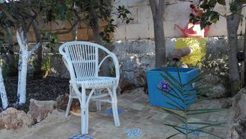 Una silla de mimbre blanca, una maceta azul con un azulejo decorativo y una zona de patio con baldosas y un muro de piedra al fondo.