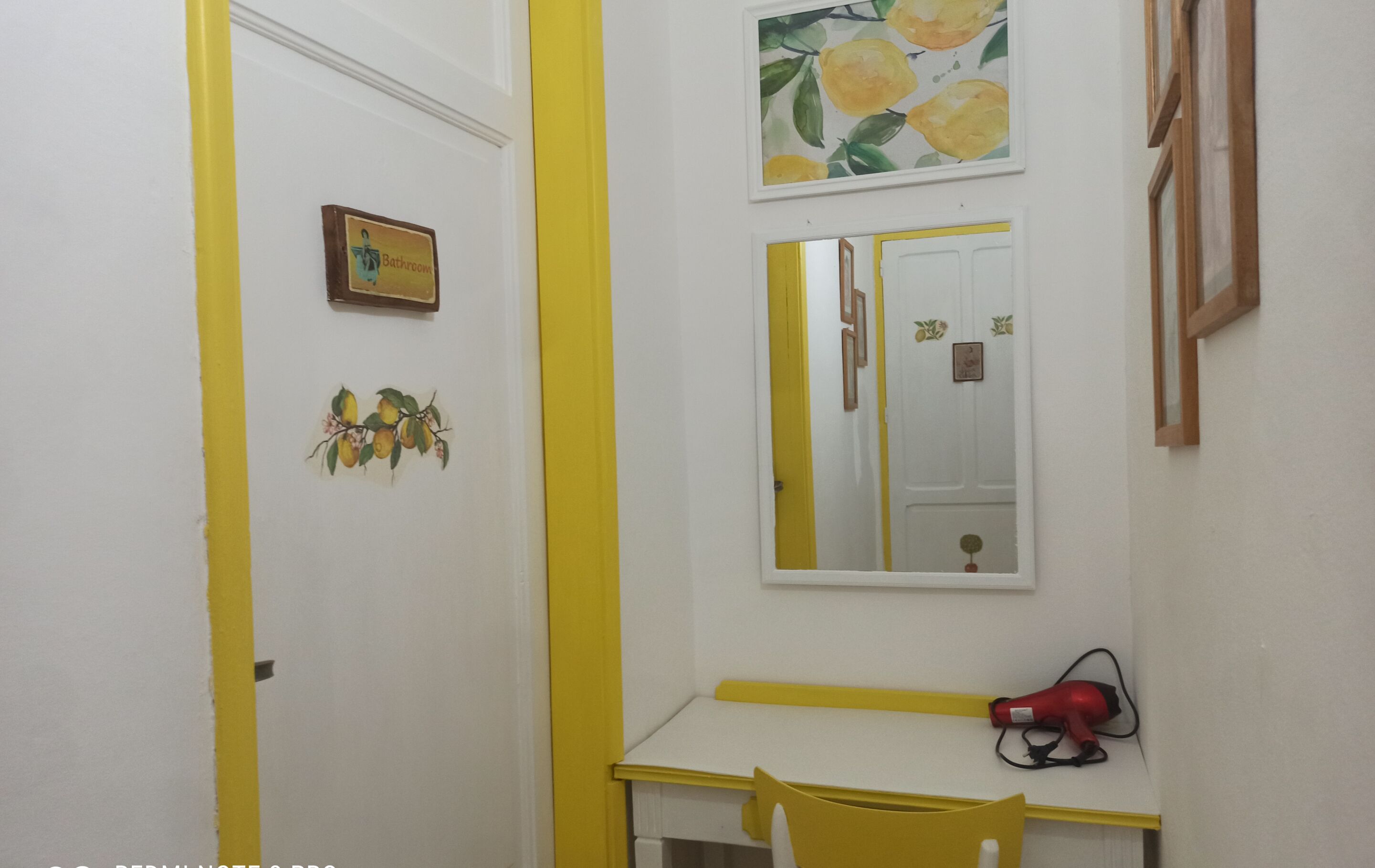 Un baño con puerta amarilla, un espejo, un cuadro enmarcado y un secador de pelo sobre una mesa blanca.