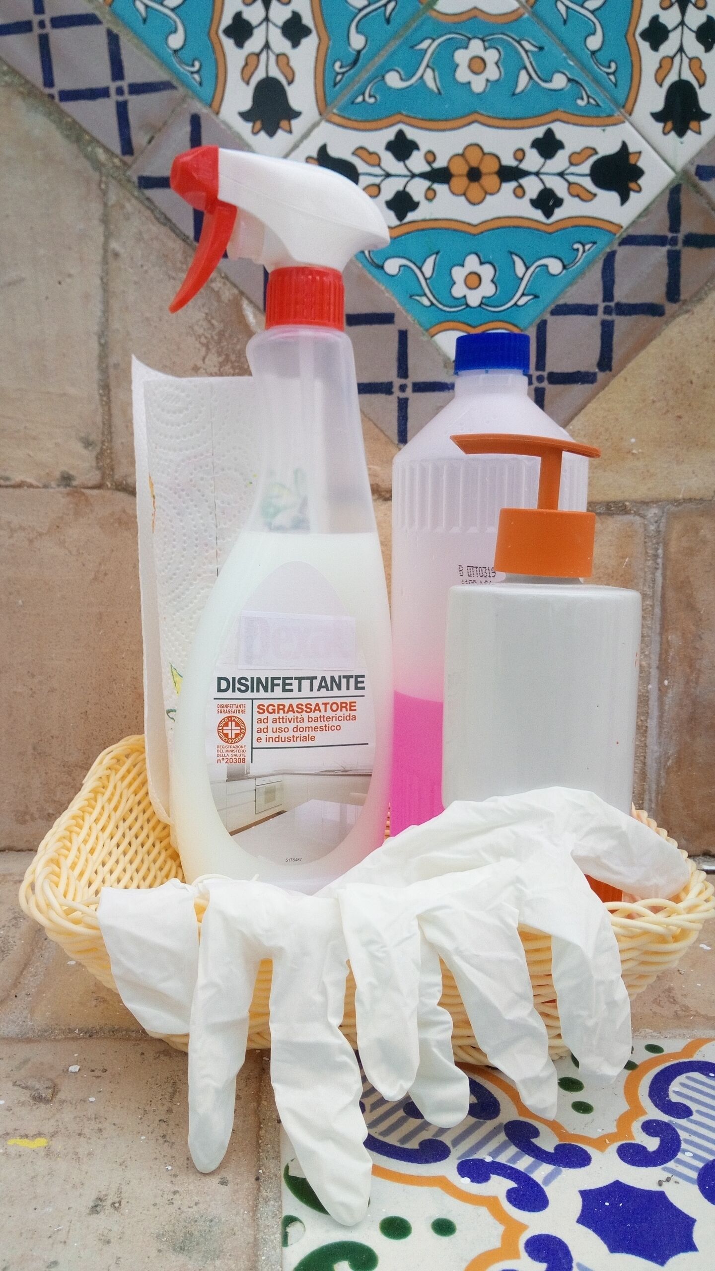 A set of cleaning supplies including a spray bottle, a bottle with a pump, and a pair of gloves, placed on a woven basket.