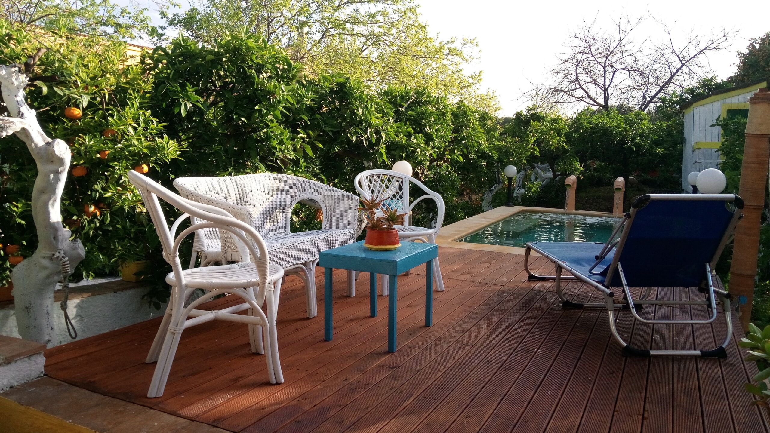 Een houten terras met rieten meubels, een kleine tafel met een plant in pot, en een zwembad.