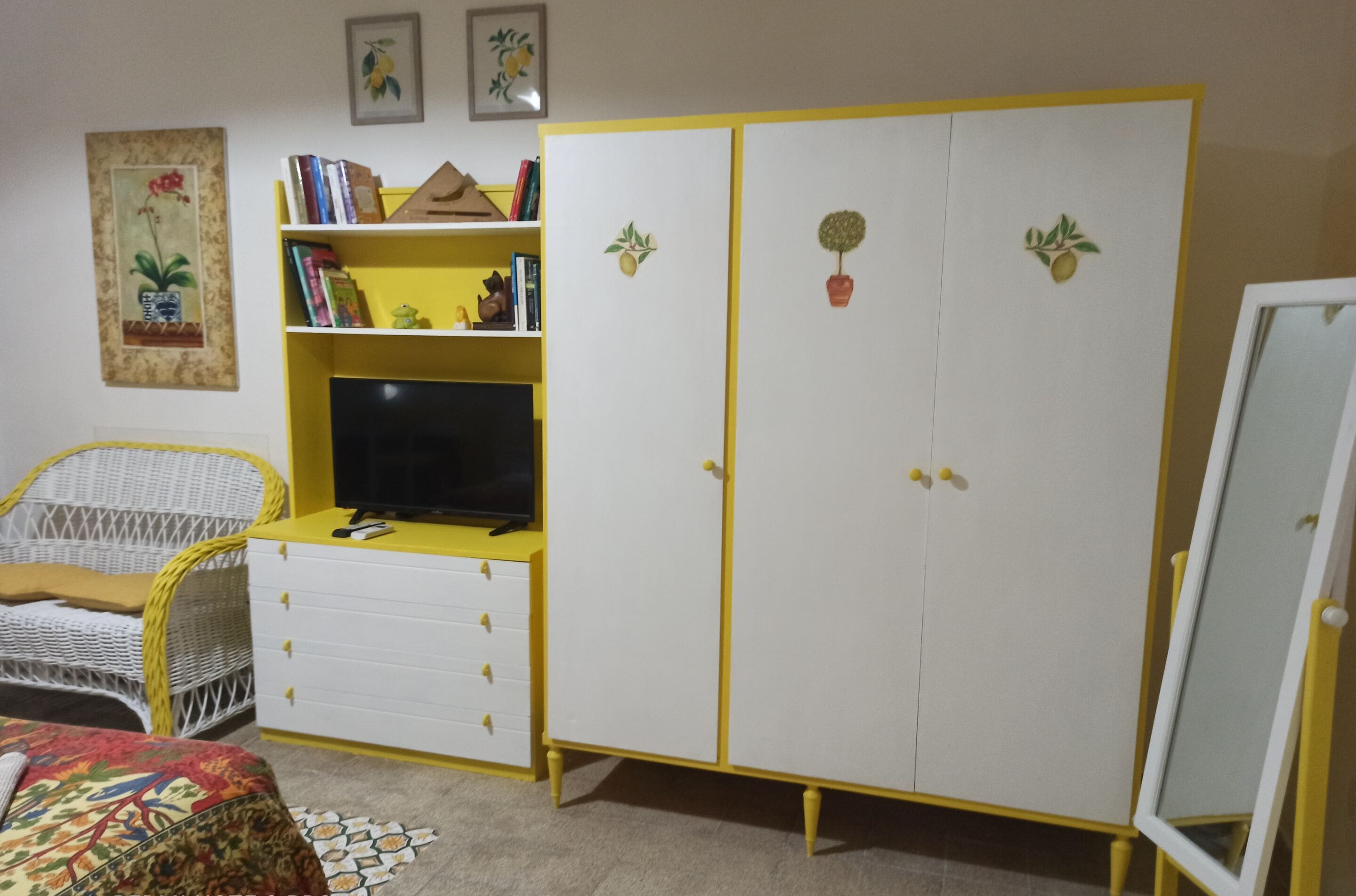A room with a white and yellow wardrobe, a television on a yellow stand, a wicker chair, and a bed with a patterned blanket.