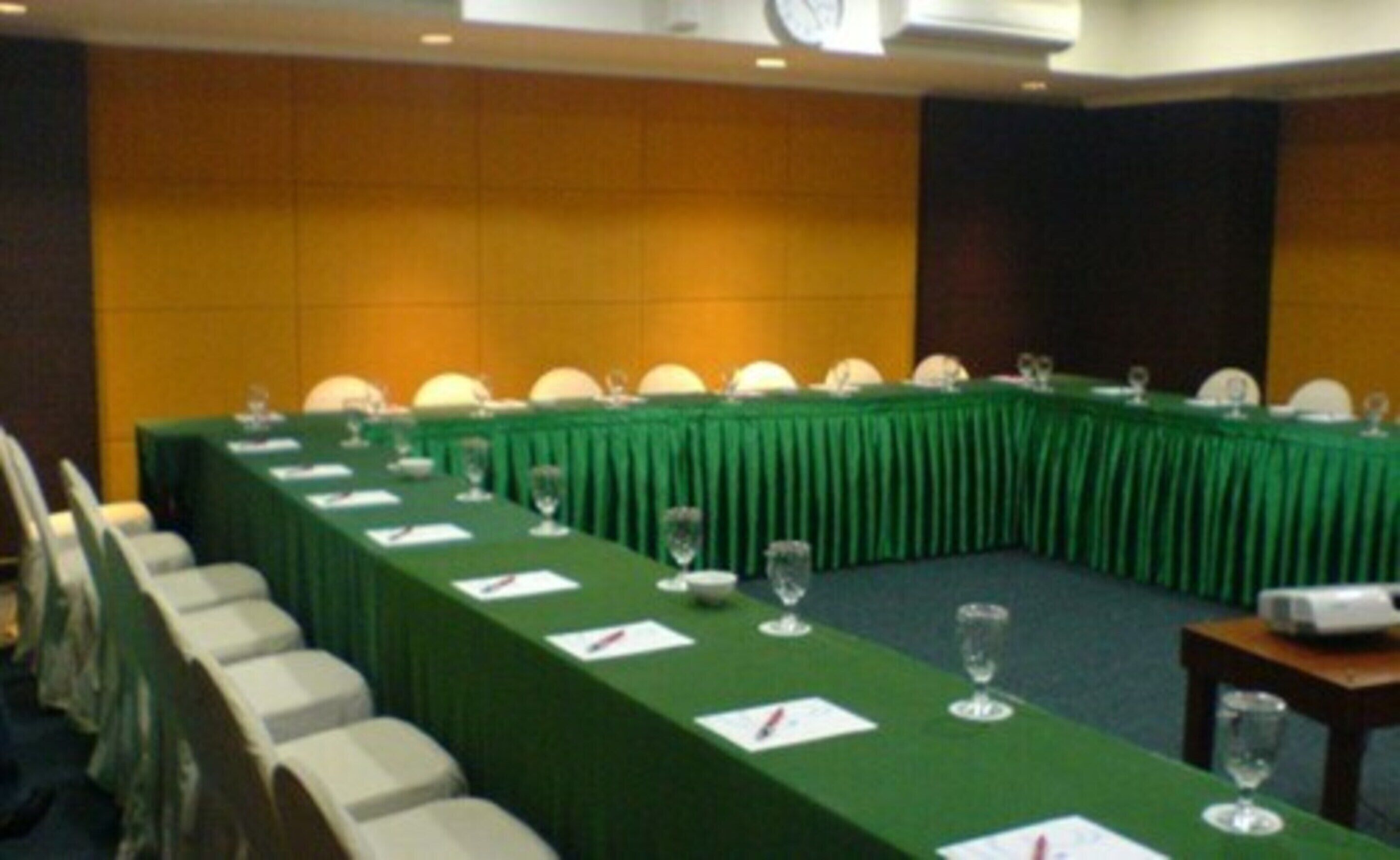Un salón de conferencias con una larga mesa cubierta con un mantel verde, sillas blancas y cristalería.