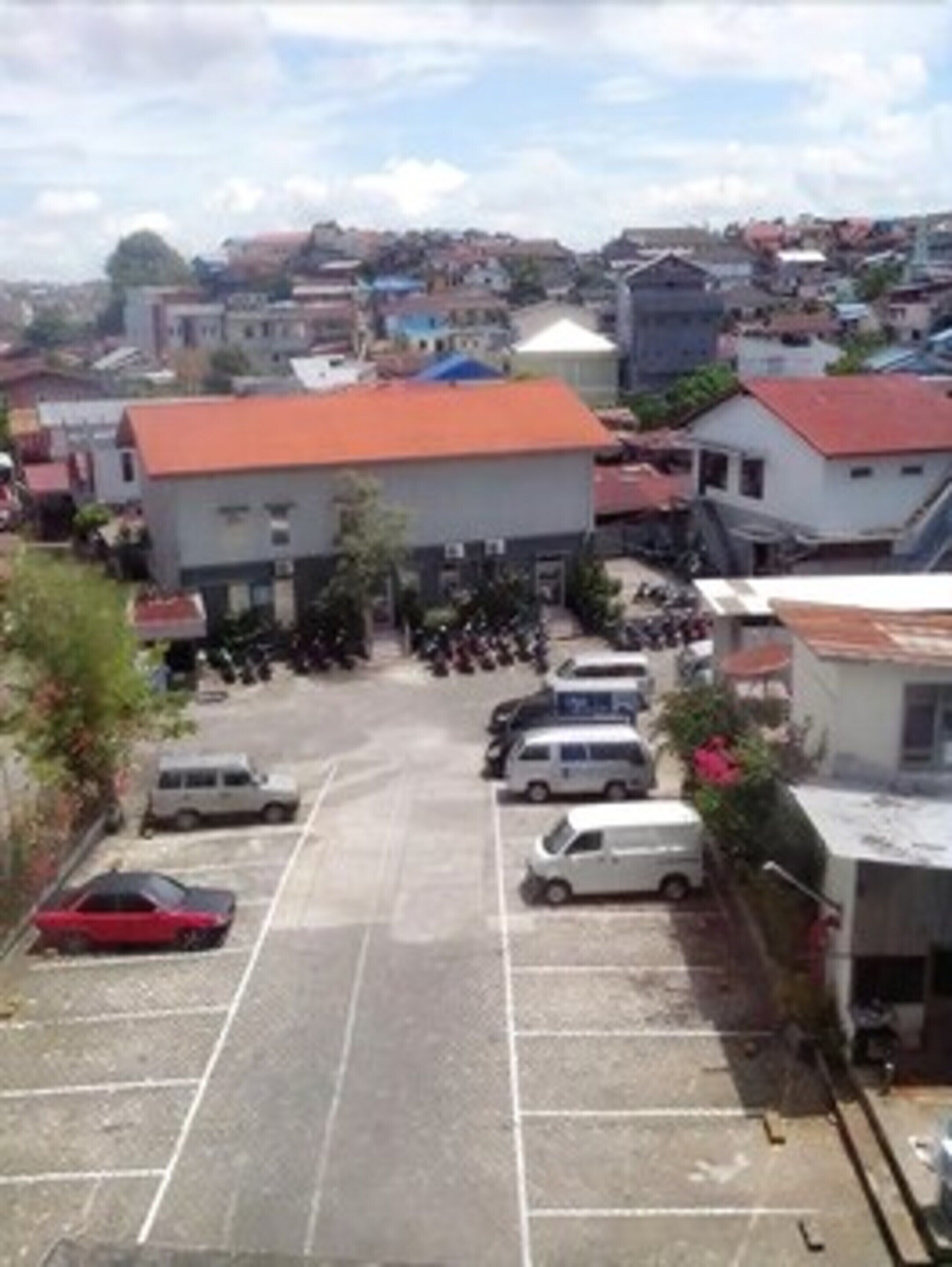 A parking lot with several cars and motorcycles, surrounded by buildings with red roofs.