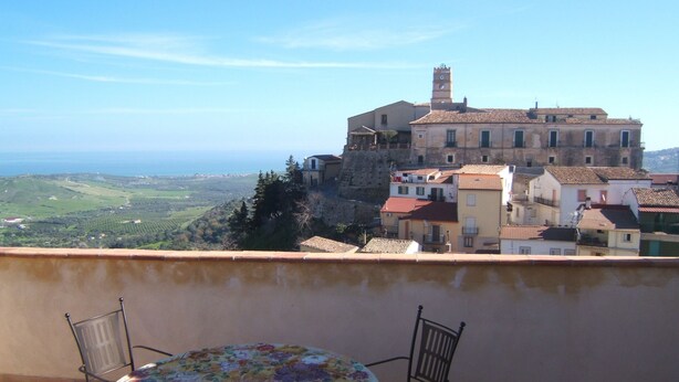 A balcony with a table and chairs, overlooking a coastal town with historic buildings and a view of the sea. - Palazzo Madeo - Residenza d'Epoca (Crosia)