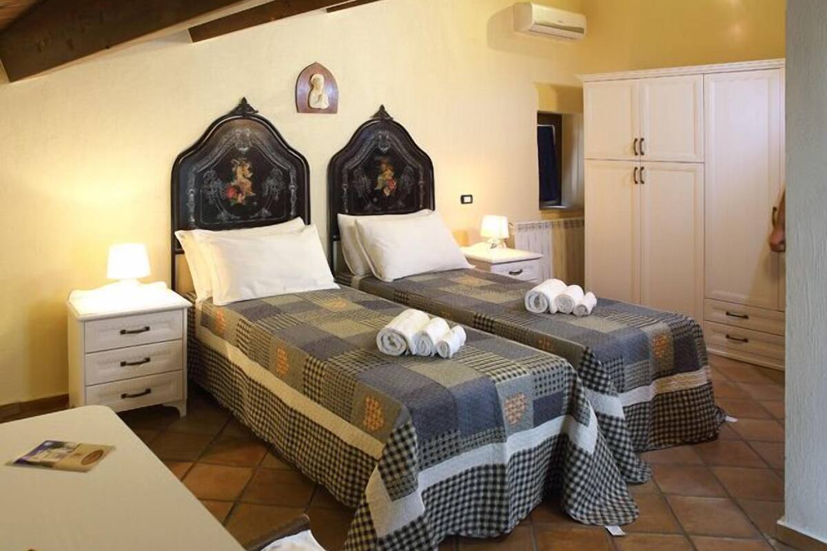 Ein Hotelzimmer mit zwei Betten, jeweils mit gesteppter Bettdecke und Kissen, einem Nachttisch mit Lampe, einem Kleiderschrank und einer Wand-Klimaanlage.