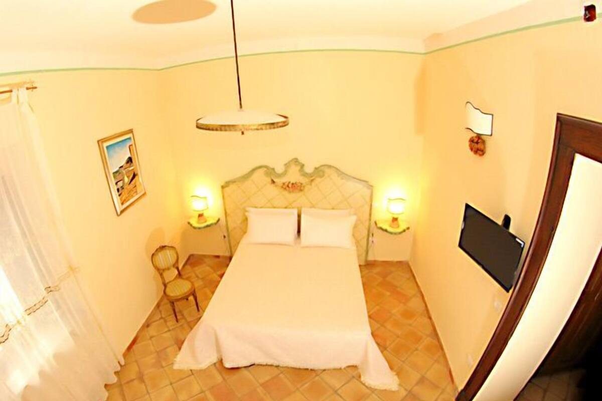 Ein Hotelzimmer mit einem Bett, zwei Nachttischlampen, einem Sessel, einem Bild an der Wand und einem an der Wand befestigten Fernseher.