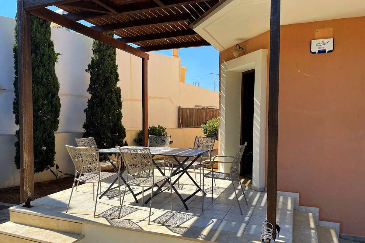 A covered outdoor patio with a table and chairs, a wooden pergola, and a security device on the wall.