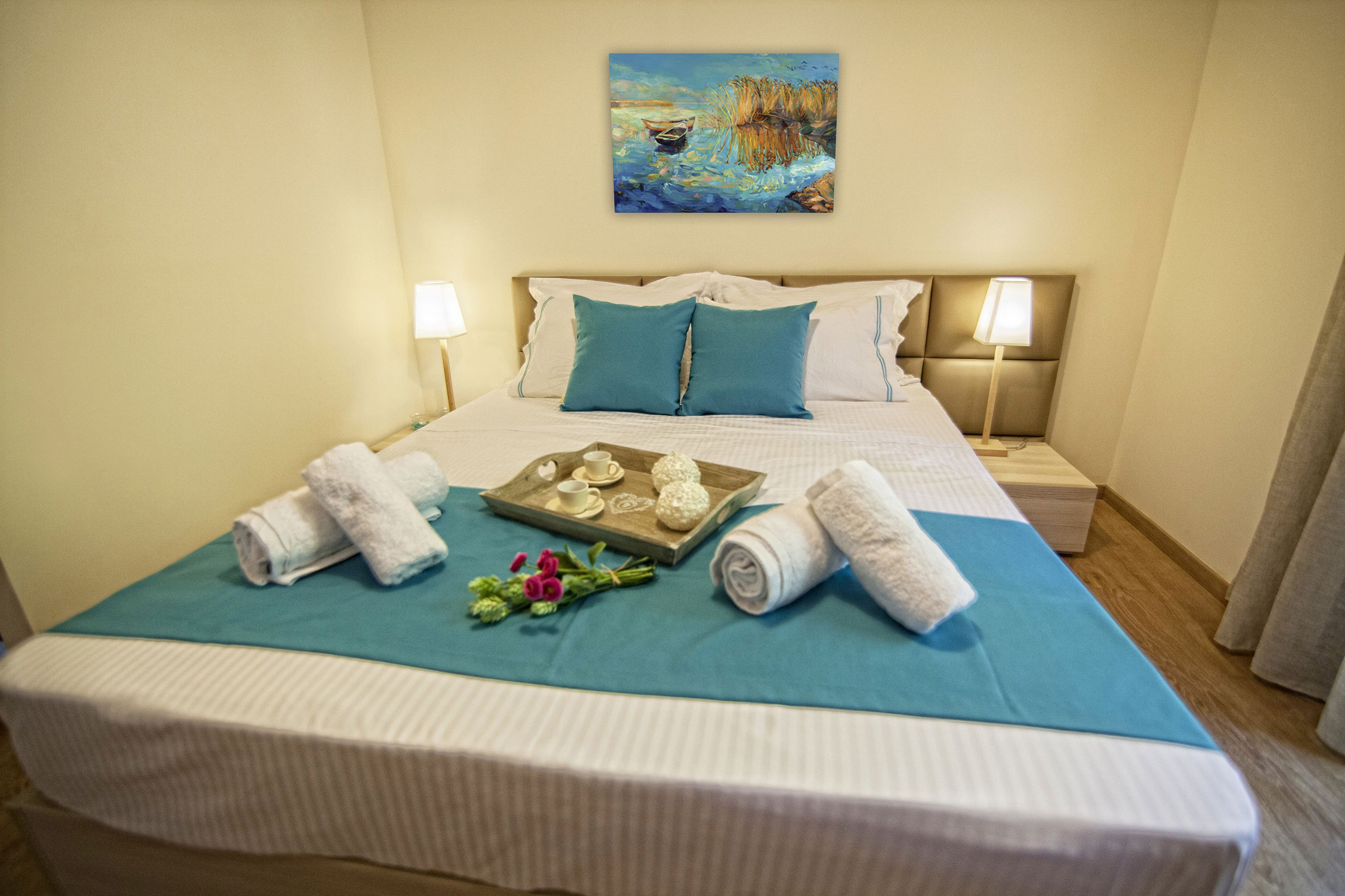 Una cama bien hecha con una colcha verde azulada, dos almohadas y una bandeja con toallas y un pequeño ramo de flores.