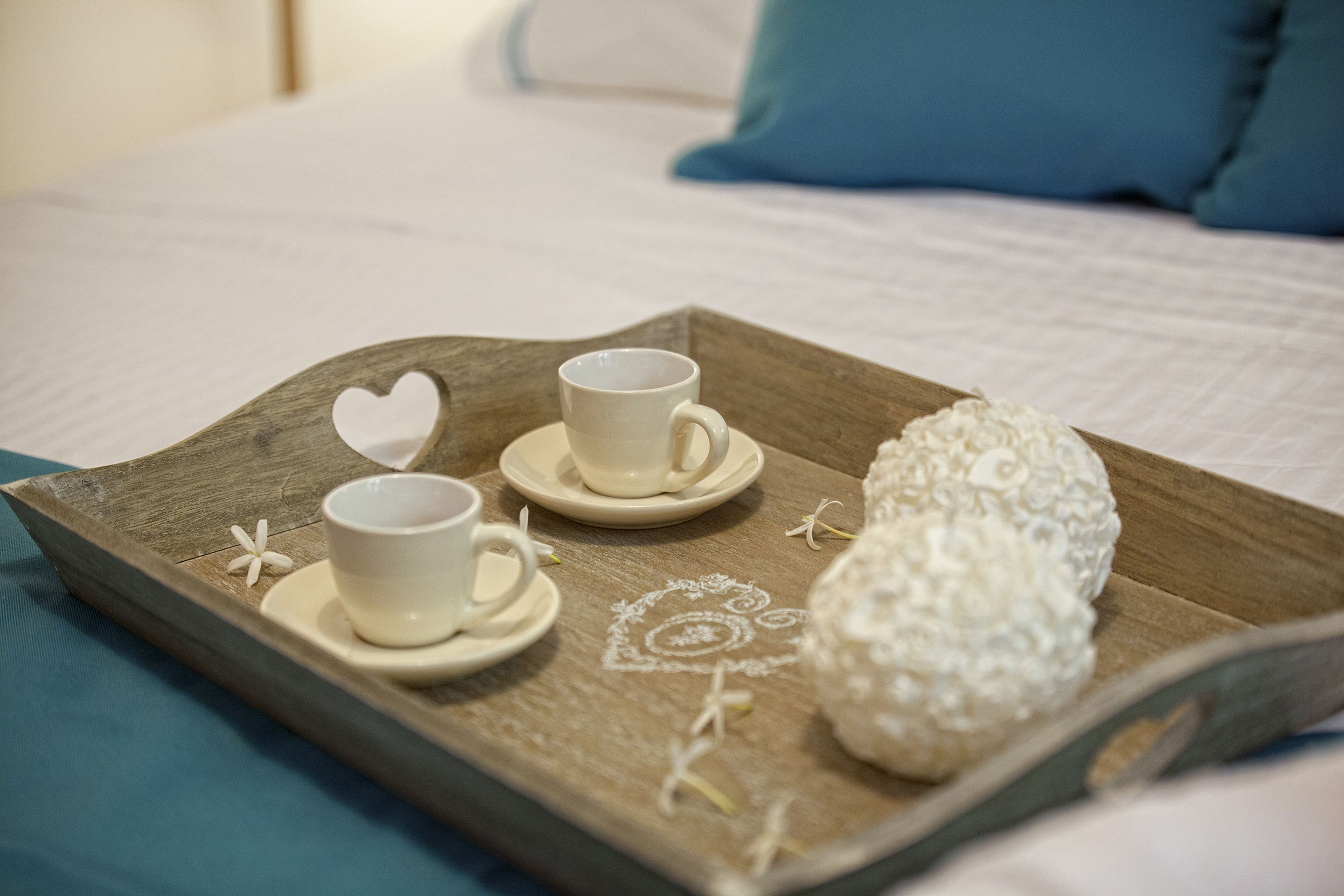 A wooden tray with two white cups and a decorative item on a bed with a blue pillow.