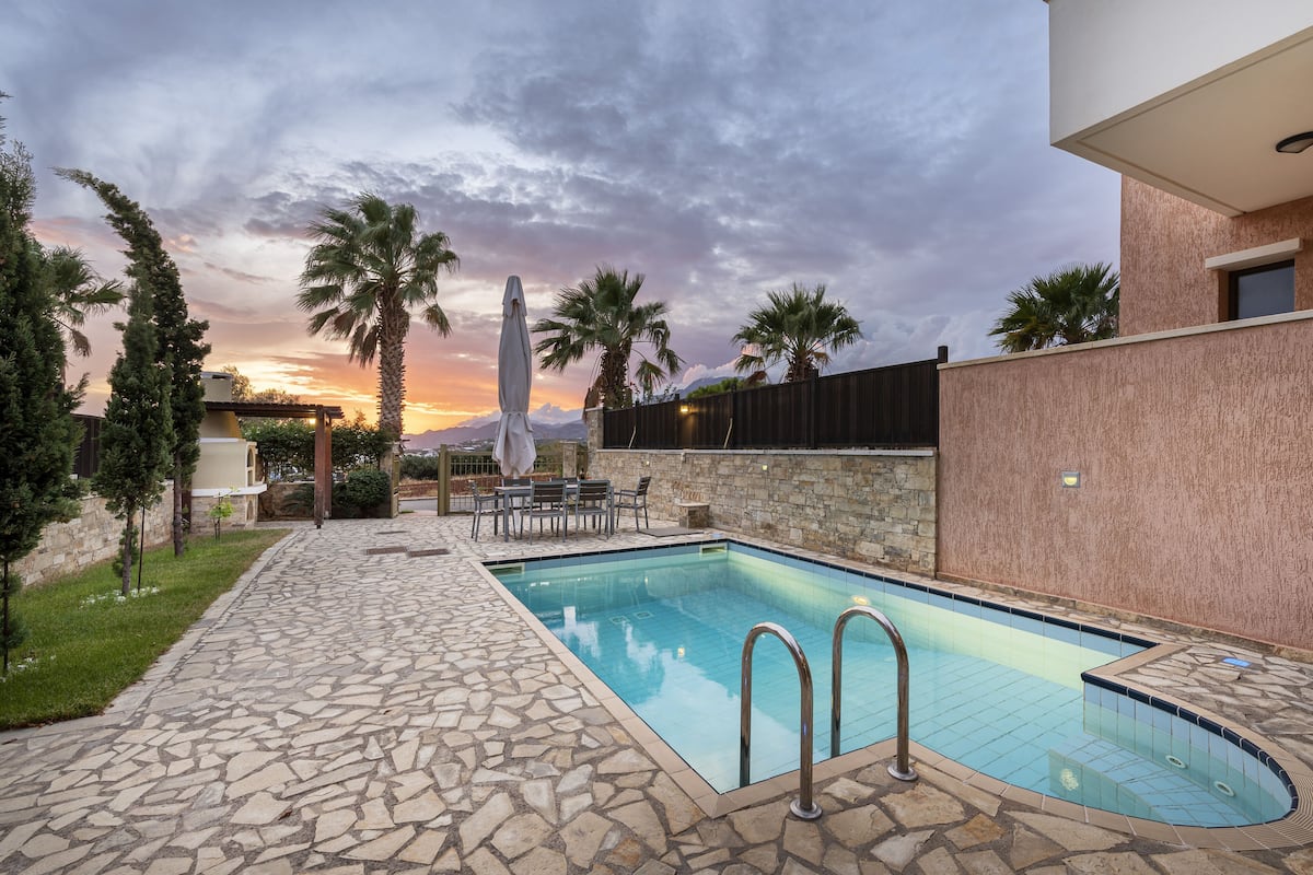 A swimming pool with stone tiles, surrounded by a stone wall and palm trees.