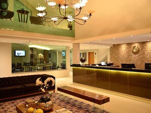 A hotel lobby with a reception desk, a sofa, a coffee table with flowers, and a television screen.