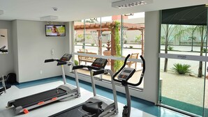 Ein Fitnessstudio mit Laufbändern, einem Fernseher und großen Fenstern mit Blick auf einen Innenhof.