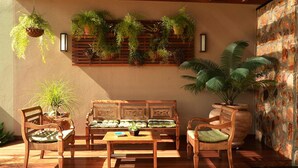 Un banc en bois avec des coussins, une table et des plantes suspendues dans une pièce ensoleillée dont un des murs est en pierre.