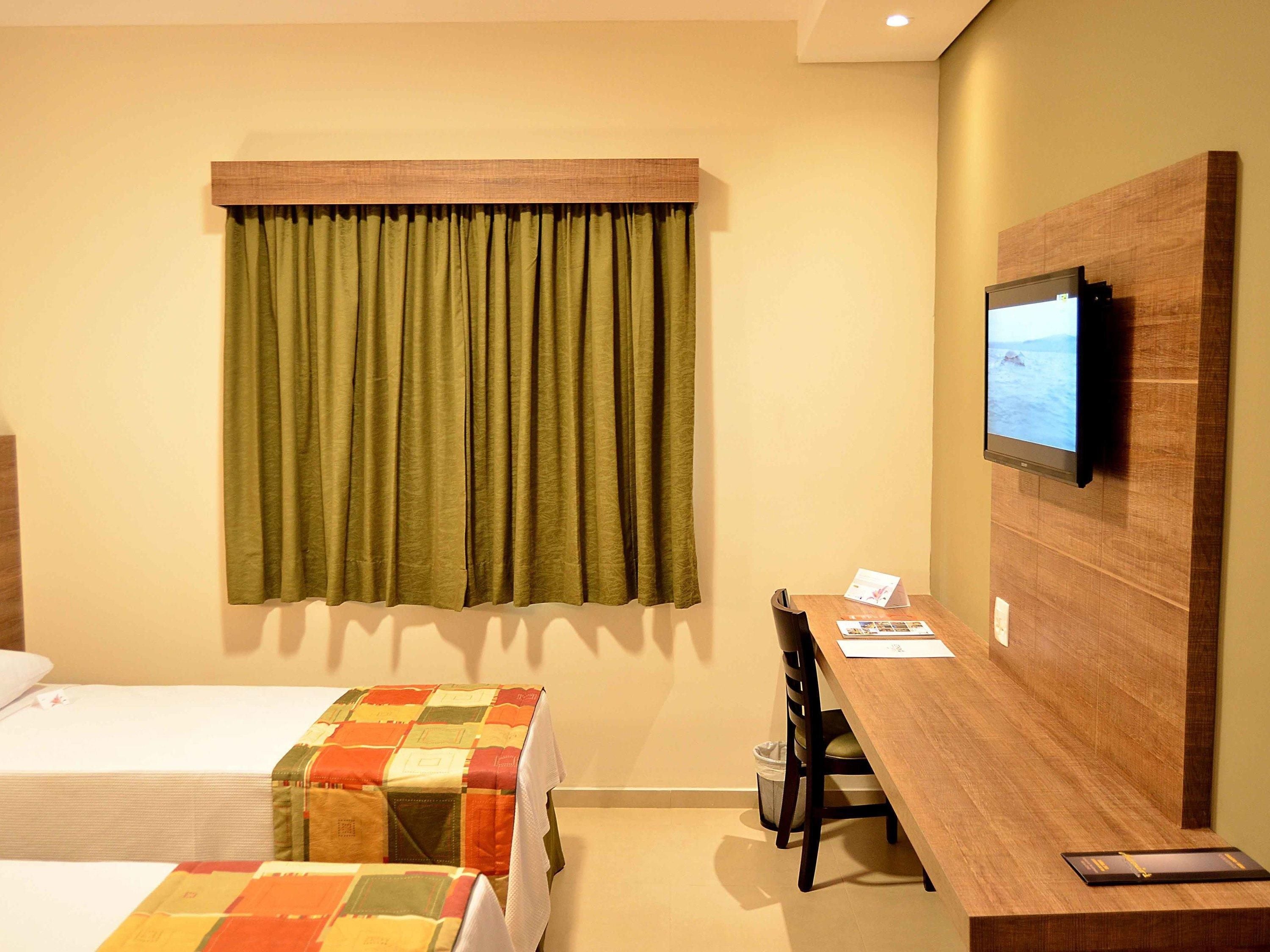 A hotel room with a bed, a desk, a chair, a television mounted on the wall, and a window with curtains.