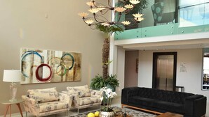 A modern living room with a black sofa, a patterned sofa, a coffee table with magazines, a floor lamp, a chandelier, and a large abstract painting.