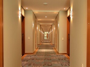 Corredor de hotel com piso em carpete, portas de madeira e luminárias fixadas na parede.