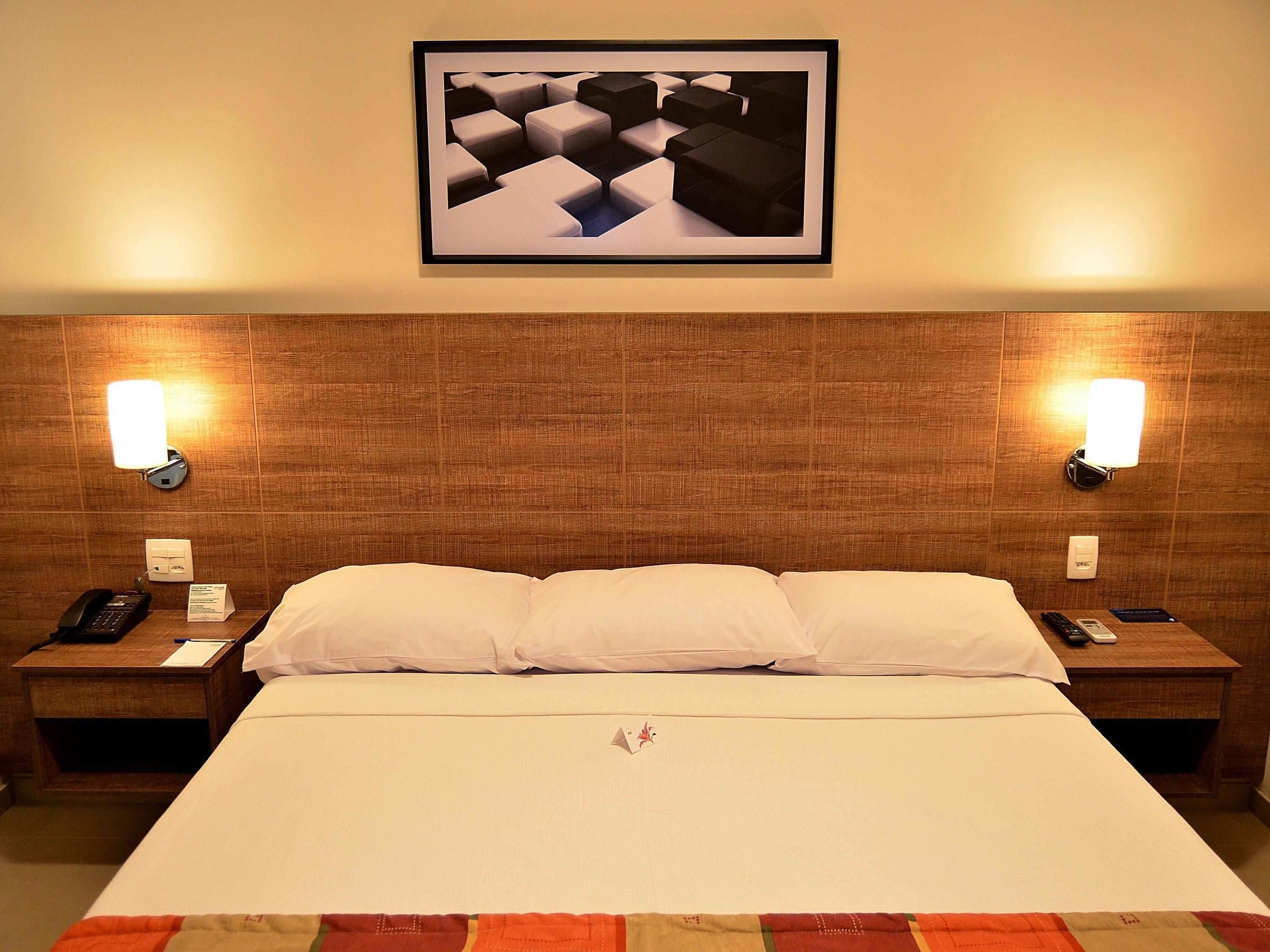 A hotel room with a wooden headboard, two bedside lamps, a framed artwork, a telephone, and a bedside table with a remote control.
