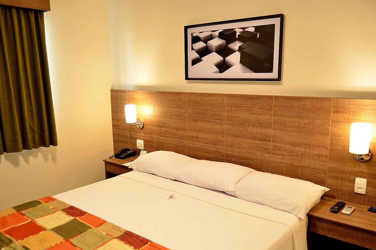 Une chambre d’hôtel avec un lit, une couette, une table de chevet, un téléphone, une télécommande et un tableau encadré au mur.