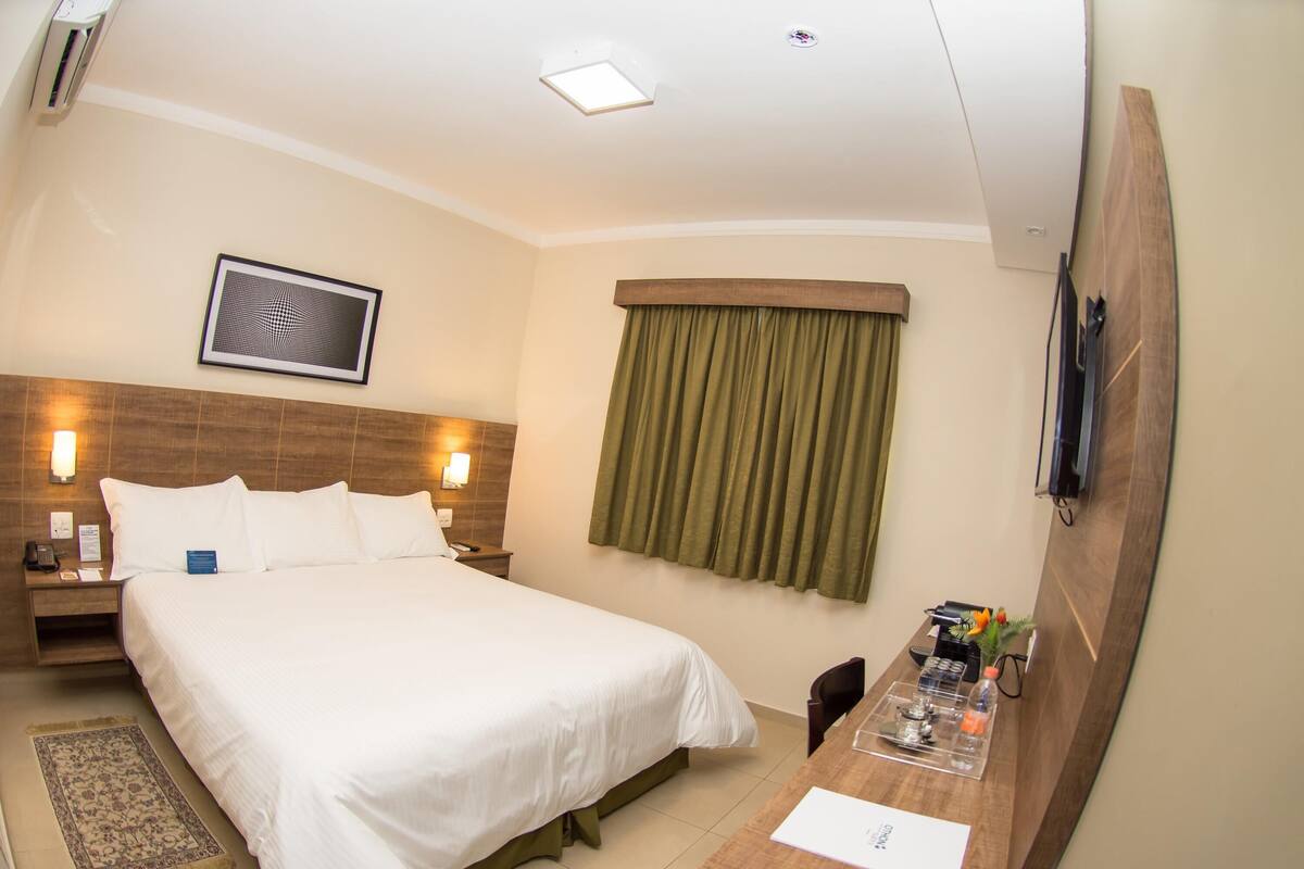 Une chambre d’hôtel comprenant un lit, une table de chevet, une lampe, un tableau, une fenêtre avec des rideaux et un bureau avec une chaise.