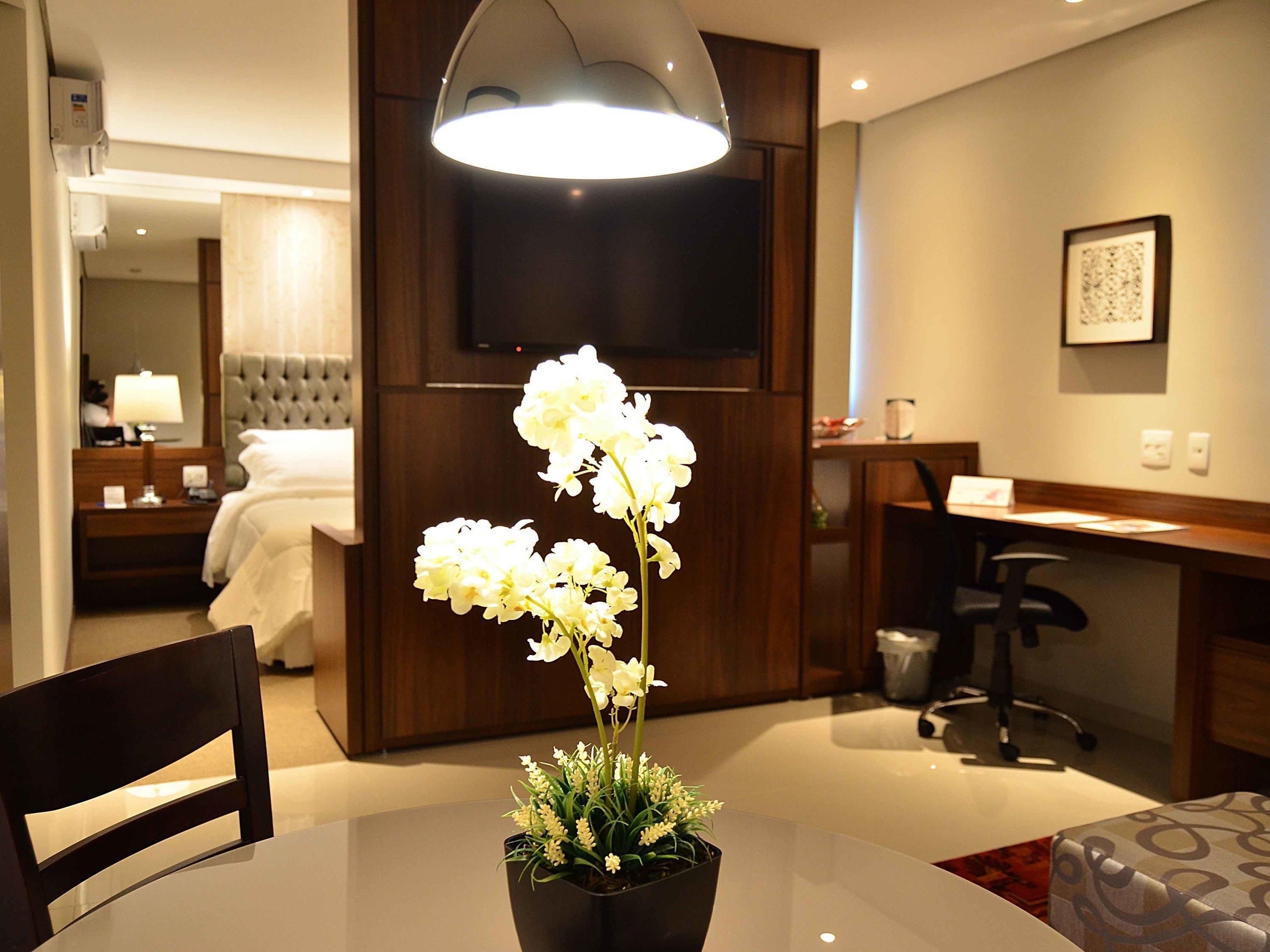 Quarto de hotel com mesa de jantar, uma orquídea em vaso, cama, televisão e escrivaninha com cadeira.