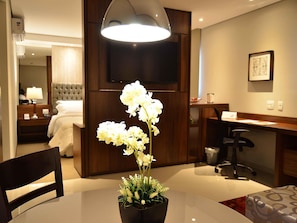 Habitación de hotel con una mesa de comedor, una orquídea en maceta, una cama, un televisor y un escritorio con una silla.