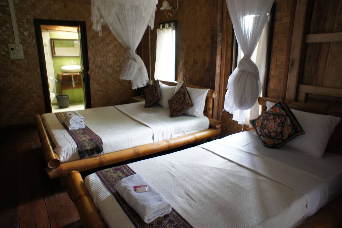 Ein Zimmer mit zwei Betten, jeweils mit weißer Bettwäsche und dekorativen Kissen. Ein Holzboden und ein Badezimmer mit einem grünen Waschbecken sind sichtbar.