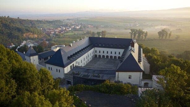 A large, historic building with multiple wings and a courtyard, surrounded by lush greenery and a rural landscape.