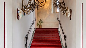 A grand staircase with red carpet, flanked by white walls and decorative antler mounts, leading to a high ceiling with a chandelier.