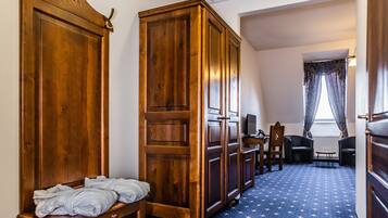 A hotel room with a blue carpet, wooden wardrobe, a bench with towels, a desk with a chair, and a window with curtains.