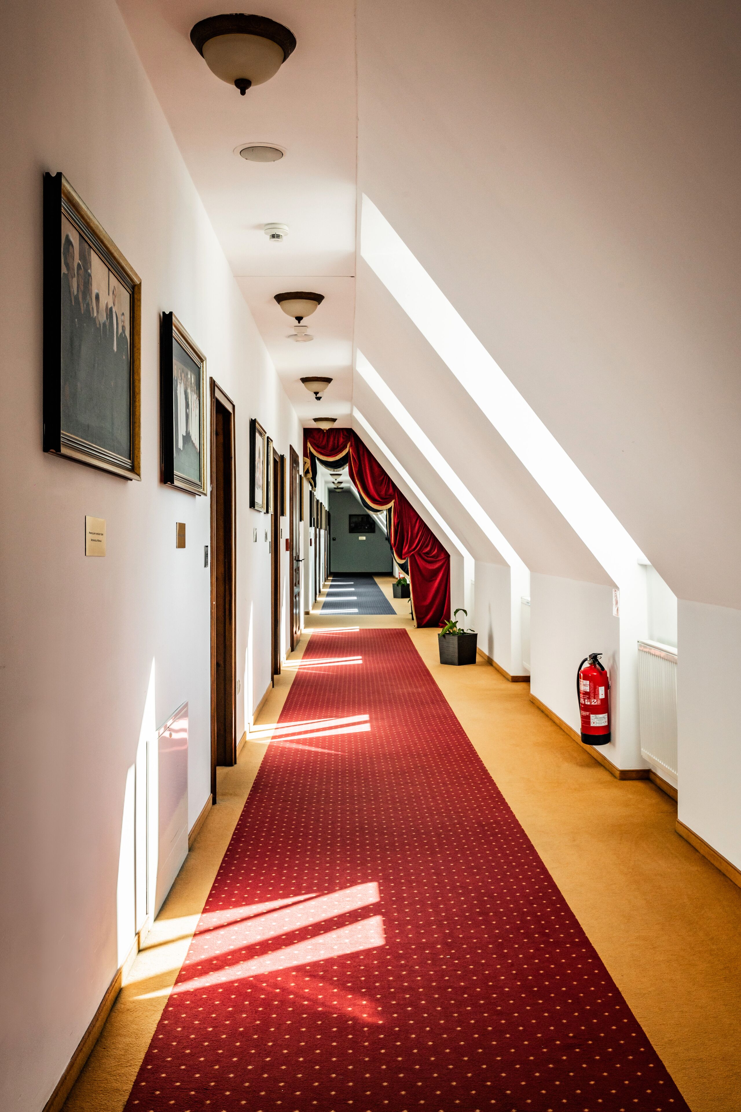 Un pasillo de hotel con alfombra roja, cuadros enmarcados en las paredes y un extintor.