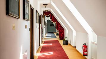 Un pasillo de hotel con alfombra roja, cuadros enmarcados en las paredes y un extintor.