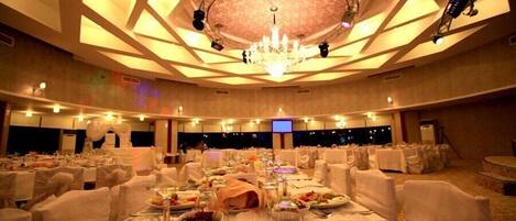 Un salón de banquetes con mesas redondas dispuestas para cenar, una araña y luces de escenario.