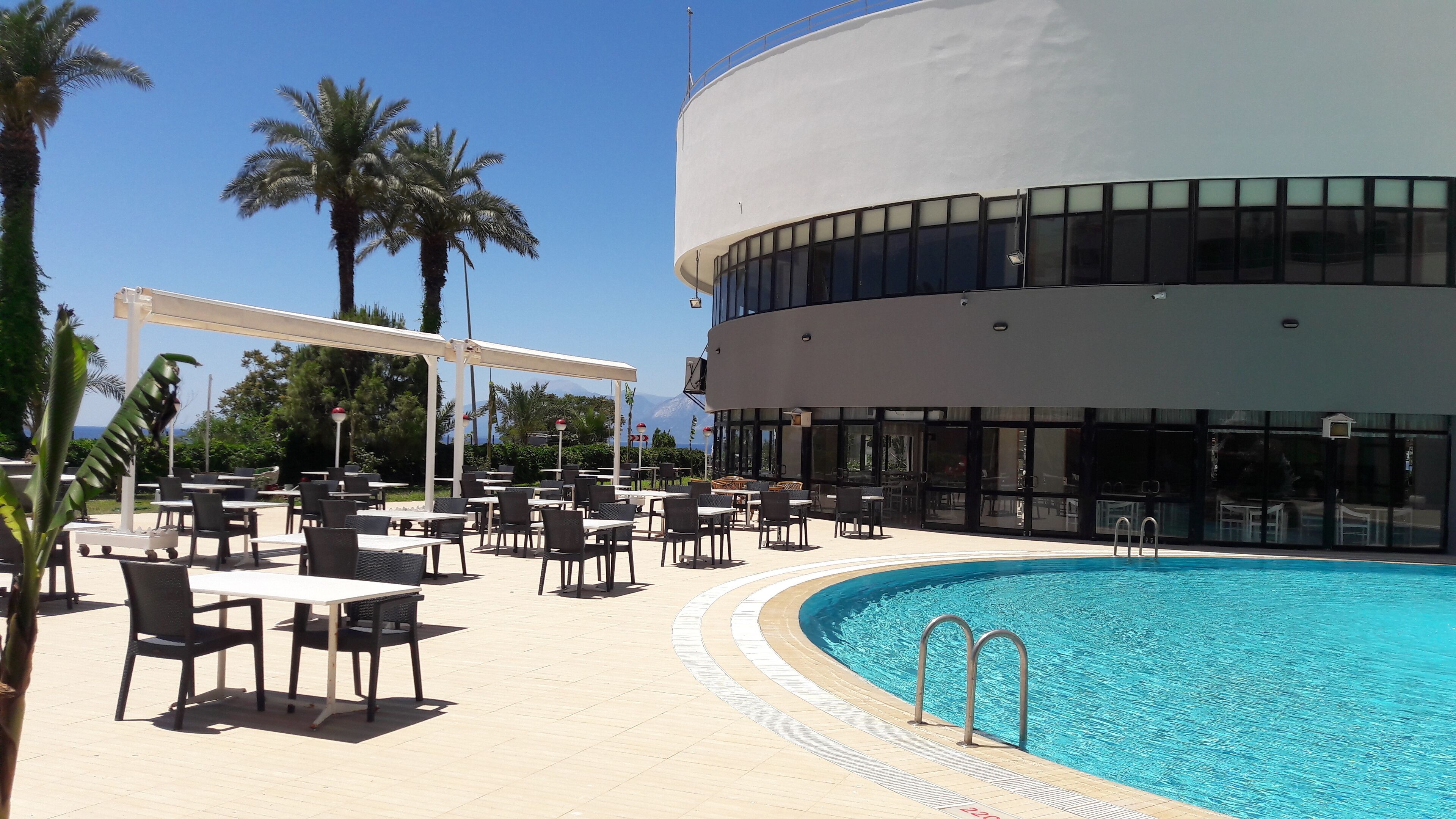 Un área junto a la piscina con mobiliario exterior, piscina y un edificio moderno con amplias ventanas.