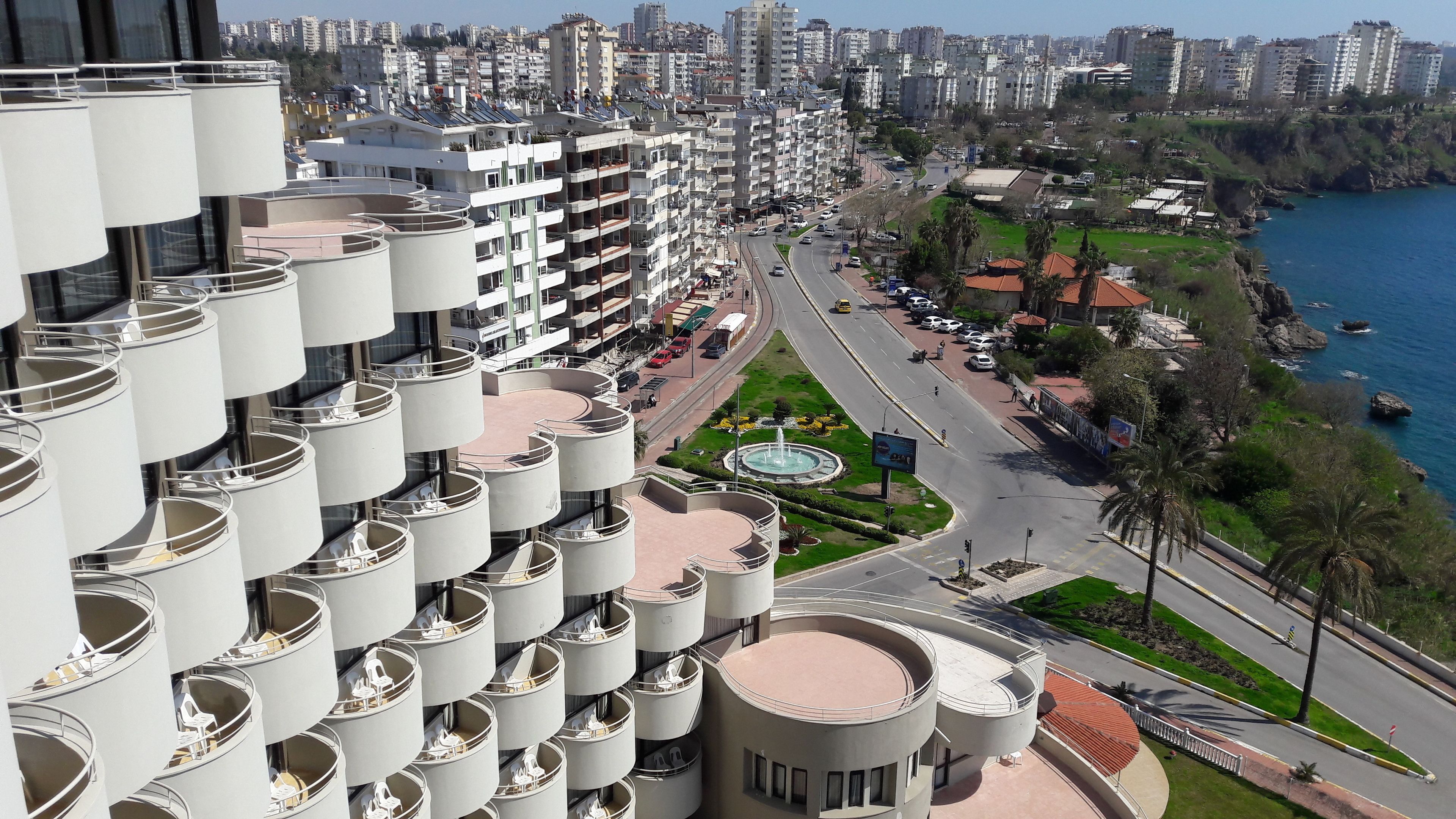 A high-rise building with multiple balconies, a central courtyard with a fountain, and a view of a coastal area with a road and parked cars.
