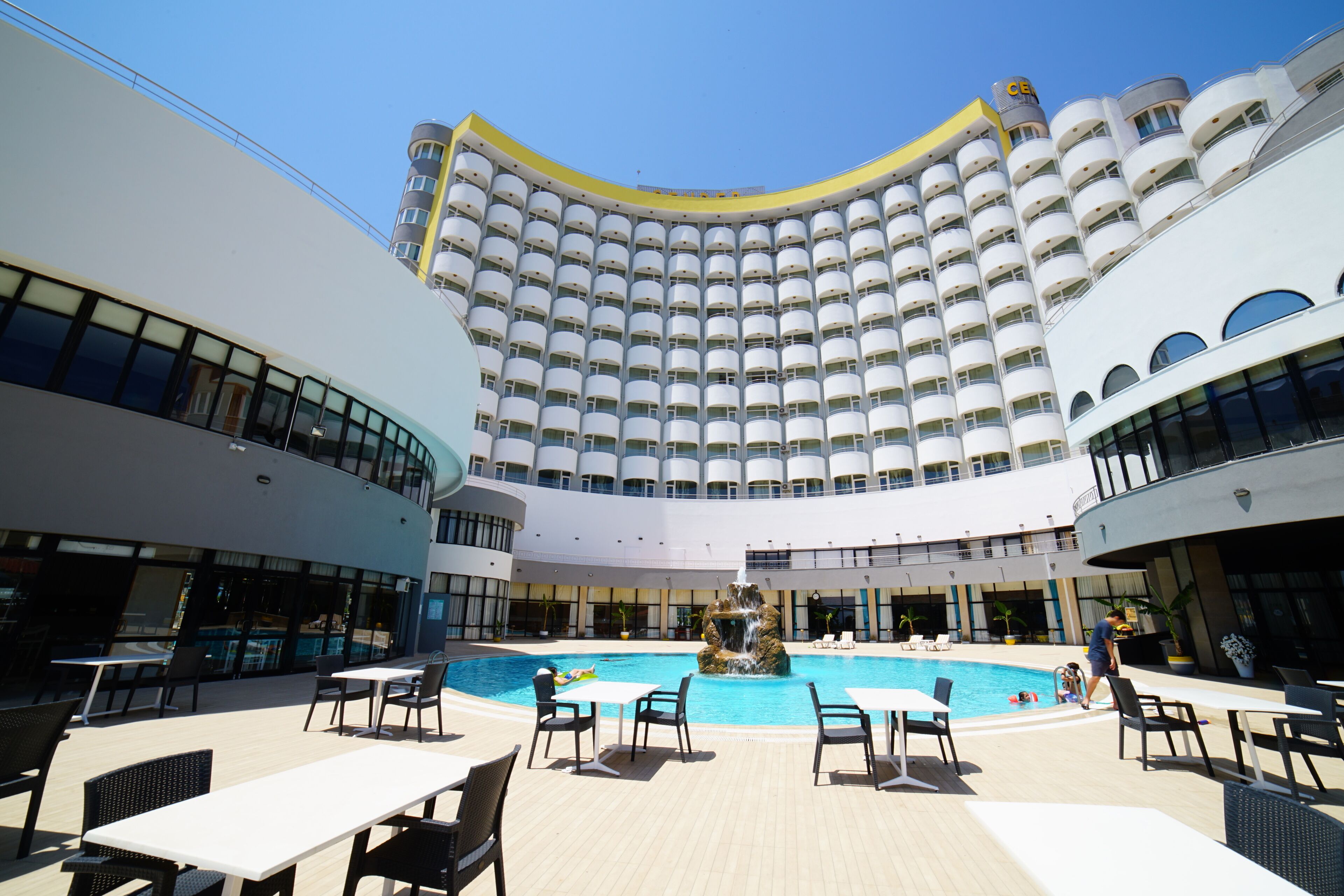 A large indoor swimming pool with multiple lounge areas and a high-rise hotel building in the background.