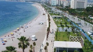 A beach with people sunbathing and walking along the shore, adjacent to a road with buildings and greenery. - Cender Hotel (Antalya)