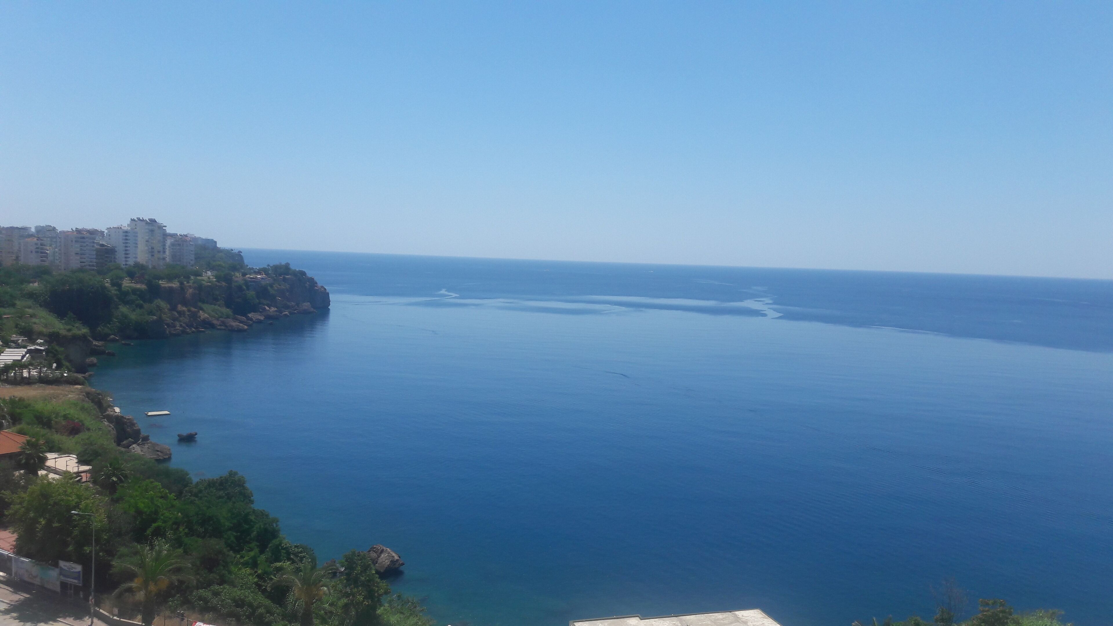 A coastal view with a clear sky, a calm sea, and a rocky coastline with buildings.
