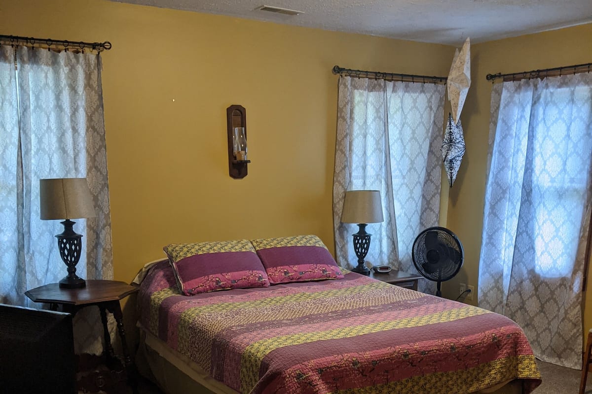 Une chambre à coucher avec un lit, deux tables de chevet avec des lampes, une horloge murale et un ventilateur de plafond.