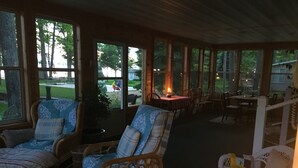 Een overdekt terras met rieten meubilair, een eettafel met stoelen en grote ramen met uitzicht op de omgeving.