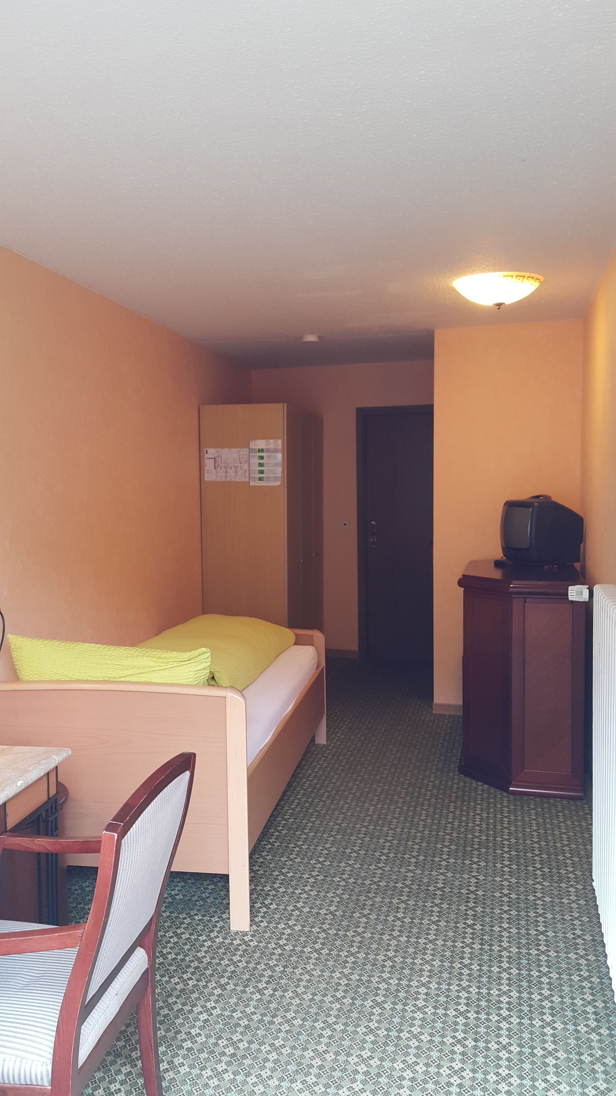 A small, single-room hotel room with a bed, a desk, a chair, a television, and a radiator.