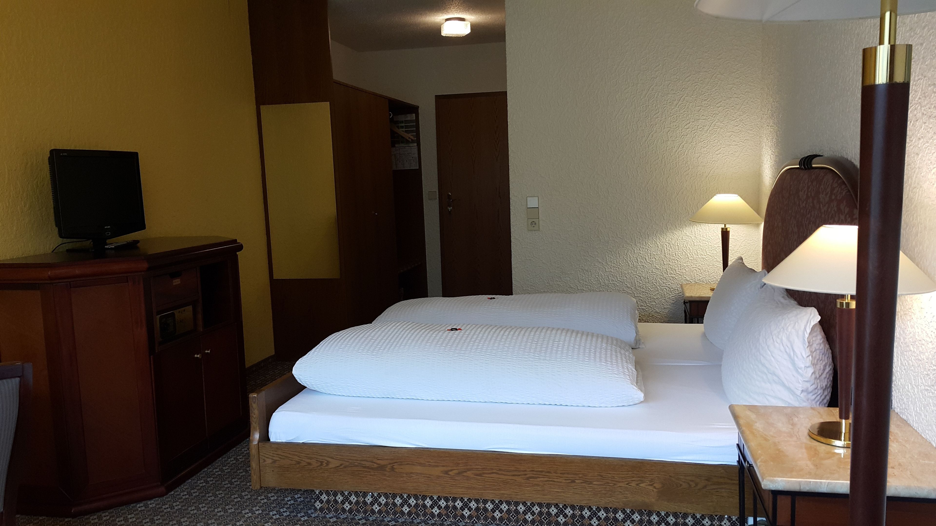 Habitació d'hotel amb llit, taulella, làmpada, televisió i armari.