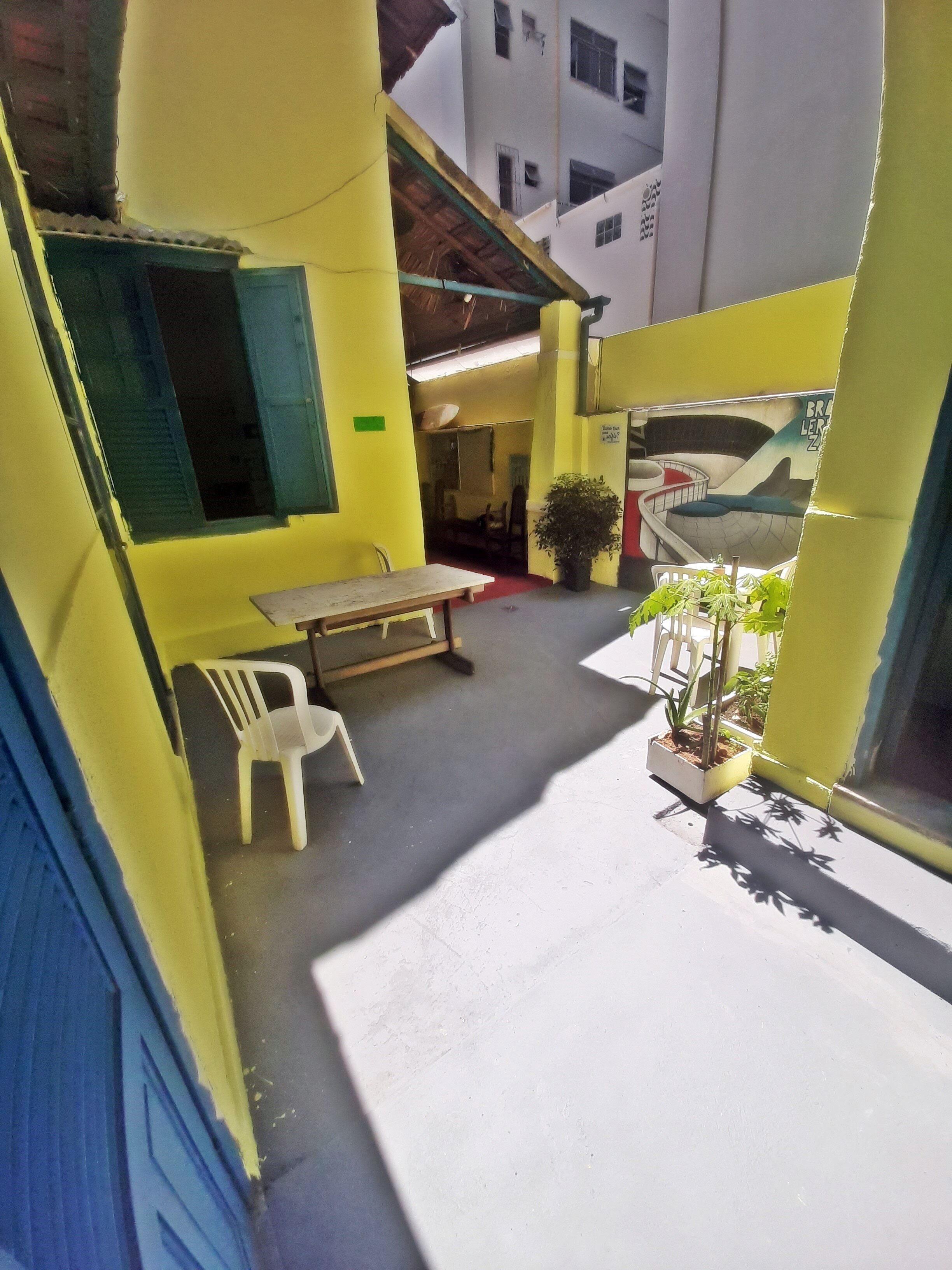 Un estrecho pasillo exterior con una mesa, sillas, plantas en macetas y un mural en la pared.
